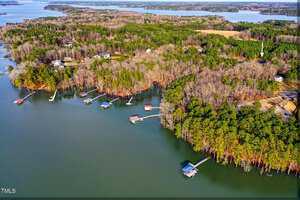 Aerial view of a lakeside landscape with dense green and brown forested areas. Several docks extend into the water, and scattered houses are visible among the trees. A large body of water surrounds the land, with more forested regions in the distance.