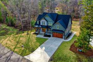 Aerial view of a two-story house with dark green siding and a stone facade. It features a wooden front porch and a two-car garage. The property is surrounded by trees and has a large driveway with a neatly manicured lawn.