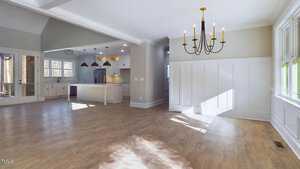 Spacious interior of an open-plan living area with light wood flooring. There is a large kitchen with an island and three pendant lights. A dining area features a black chandelier, and multiple windows allow natural light to flood the space.