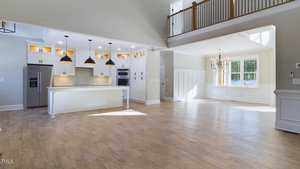 Spacious, modern open-plan kitchen and living area with light wood flooring. Features include a large island with white cabinets, three black pendant lights, stainless steel appliances, and a dining area with a window and chandelier.