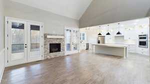 Spacious open-concept room with hardwood floors, featuring a kitchen with white cabinets and a large island with pendant lights. A stone fireplace is on the left with doors leading to a balcony. Large windows provide natural light.