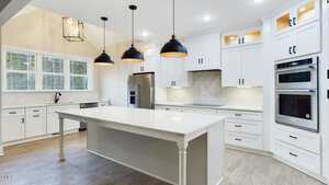 A modern kitchen with white cabinets and a large central island featuring a light countertop. Three black pendant lights hang above the island. Stainless steel appliances, including a fridge and double oven, are set against a backsplash.