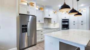 Modern kitchen with white cabinets, stainless steel refrigerator, built-in oven and microwave, and an electric cooktop. An island with a white quartz countertop and three pendant lights is in the foreground. The floor is light wood.