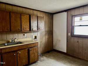 A small, empty kitchen with wood-paneled walls and a tiled floor. There is a window on the right, wooden cabinets above and below a countertop on the left, with a sink and space for appliances. The corner shows signs of water damage.