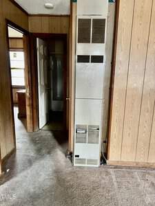 A narrow, wood-paneled hallway features a tall, white wall heater in the center. Open doors on either side lead to other rooms. The carpeted floor appears worn and the walls show signs of age.