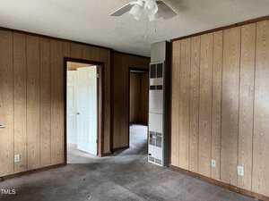 A room with wood-paneled walls and a ceiling fan. There is a doorway leading to another room with an open white door. The carpet is worn and the room appears empty. A vertical metal heater is visible in one corner.