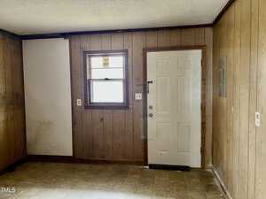 Room with wood-paneled walls and a textured ceiling. A window with blinds is on the left, above a small section of white wall. A closed white door with a mail slot is next to the window. The floor has a tan linoleum covering.
