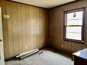 A small, empty room with wood-paneled walls and a single window. The carpeted floor shows some debris, including wooden planks and tools. The ceiling is off-white, and there is a wooden dresser partially visible on the right.