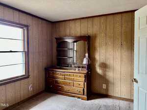 A small room with wood-paneled walls and a beige carpet. A dresser with multiple drawers stands against the wall, topped with a mirror and a small pink lamp. A window is on the left, and a white door is partially open on the right.