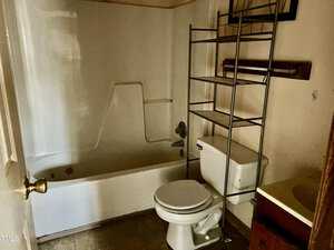 A small, dimly lit bathroom with a white bathtub, a toilet, and a metal shelving unit above the toilet. The floor appears to be tiled, and there's a sink with a brown cabinet to the right. A white door is partially open.