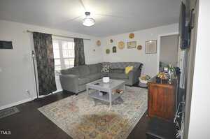 Living room with a grey sectional sofa and patterned cushions. A large rug covers the dark wood floor. The walls have decorative plates and a hanging picture. A coffee table is at the center, and a TV is mounted on a wall near a window with dark curtains.