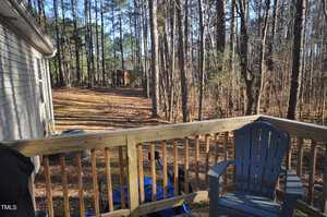 View from a deck with a wooden railing overlooking a backyard with tall trees. A gray plastic chair is on the deck, and a blue tarp covers an object. A small wooden structure is visible in the distance among the trees.