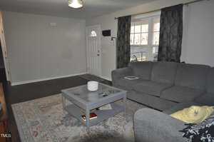 Living room with gray walls and dark wood floor. A gray sectional sofa and coffee table are placed on a patterned area rug. A large window with dark curtains faces the sofa. A small ceiling light hangs near the entrance door.