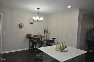 A modern dining area features a table with two chairs, a hanging light fixture, and an exercise bike against the wall. There are decorative items on a small wall shelf and a kitchen island in the foreground. A person is partially visible to the right.