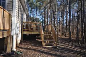 A wooden deck with a staircase leads to the side of a house. The house is in a wooded area, surrounded by leafless trees. The ground is covered with brown leaves and some patches of grass. The sky is clear, and sunlight filters through the trees.