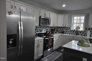 Modern kitchen with white cabinets and dark countertops. Stainless steel refrigerator and oven are present. The backsplash features a geometric pattern. A small island holds green glassware, and a window allows in natural light.