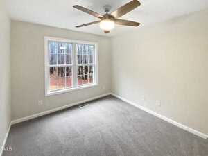 A neutral-toned, empty room with light beige walls and a beige carpet. A ceiling fan with a light fixture is centered above. A single window on the far wall reveals a view of bare trees and ground outside.
