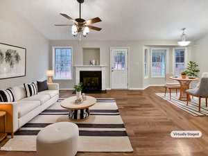A virtually staged living room with light wood flooring and neutral walls. It features a white sofa, striped rug, circular coffee table, and a white ottoman. Nearby, a dining area includes a round table and chairs. A fireplace and ceiling fan are present.