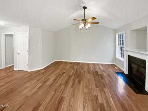A spacious, empty living room with light wooden floors and light gray walls. A ceiling fan with lights is centered above. There's a white door on the left, a window on the right, and a fireplace with a white mantel on the right wall.