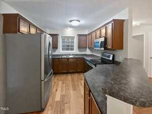 A kitchen with wooden cabinets and a dark stone countertop. It includes stainless steel appliances such as a refrigerator, stove, and microwave. A window is centered above the sink, and the kitchen flooring is wooden.