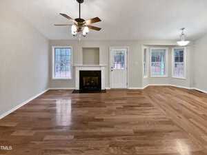 A spacious living room with wooden floors, a ceiling fan, and a fireplace. A white door and two windows are on the back wall. To the right, a semi-open area with three bay windows features a hanging light fixture. Walls are painted light gray.