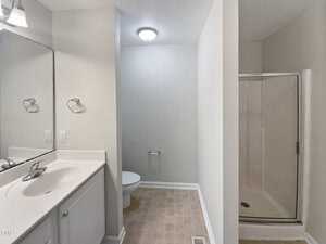 A bathroom with beige tile flooring features a white vanity with a sink and mirror on the left, a toilet visible against a light gray wall, and a glass-enclosed shower on the right. A ceiling light fixture illuminates the space.