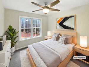 A bedroom with a light wooden bed frame and white bedding. Two bedside tables with lamps are on either side of the bed. A large window lets in natural light. A potted plant is beside a dresser. The room features a ceiling fan and abstract artwork.