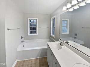 A bathroom featuring a white bathtub with a shower fixture, a large mirror above a double-sink vanity with white cabinetry, and two windows. The floor has light-colored tiles, and the walls are painted in a soft neutral tone.