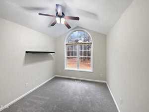 A small, empty room with a vaulted ceiling, featuring a large arched window and a simple black wall-mounted shelf. The walls are off-white, and the floor is carpeted in gray. A ceiling fan with lights is centered on the ceiling.
