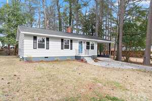 The single-story house at 320 Zollicoffer Avenue, Henderson, features white siding and black shutters, nestled among trees. A small porch with a blue front door graces the entrance, while a sloped front yard and concrete driveway complete the welcoming scene under the clear blue sky.
