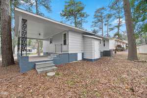 Single-story white house with a blue foundation, surrounded by pine trees. The house has a covered porch with black decorative supports and concrete steps leading to it. The yard is covered with fallen leaves. A neighboring house is visible in the background.
