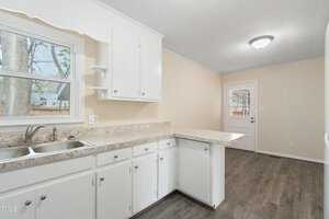 A kitchen with white cabinets, a dual sink, and a light granite countertop. The floor is dark wood, and there is a window above the sink. A door with a window is in the background, leading to another room.