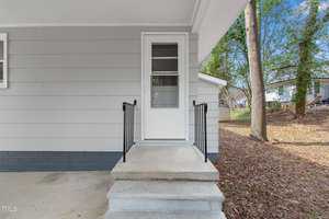 A small concrete porch with three steps leads to a white door on a light gray house. Black metal railings are on either side of the steps. The ground is covered with fallen leaves, and a tree trunk is visible on the right.