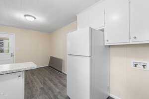 A kitchen with light-colored walls and dark flooring features a white refrigerator and white cabinetry. A small section of countertop is visible. A closed door with a window is on the left side of the room.