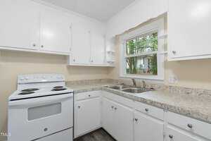 A small kitchen featuring white cabinets, a marble-effect countertop, a white electric stove with four burners, a double stainless steel sink, and a window above the sink. The floor is dark wood, and the walls are light beige.