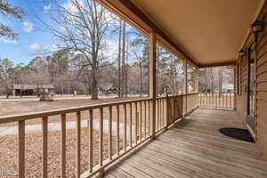 A wooden porch with railings extends along the side of a house, overlooking a yard with bare trees. A pathway leads to other wooden structures in the background under a partly cloudy sky.