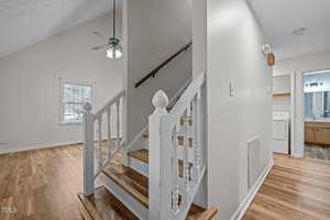 A small staircase with white balusters and wooden steps leads to an upper level. The foreground shows a room with wood flooring. A ceiling fan is visible above. In the background, there is a laundry area with a washing machine and a bathroom.