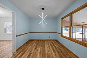 An empty room with light blue walls and wooden floor. A modern pendant light hangs from the ceiling. A large window on the right shows an outdoor area. An adjacent room with similar flooring is visible through an open doorway.