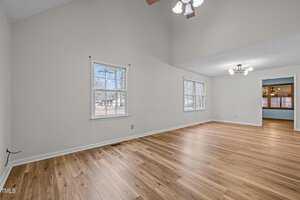 A spacious, unfurnished room with light wood flooring and cream walls. Two ceiling fixtures provide lighting. Two windows on the left wall let in natural light. An open doorway leads to another room with darker flooring.