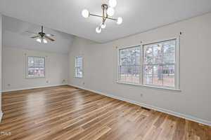 A spacious, empty room with light wood laminate flooring and cream-colored walls. Two windows on the right let in natural light, showing trees outside. The ceiling features two different light fixtures: a modern chandelier and a ceiling fan.