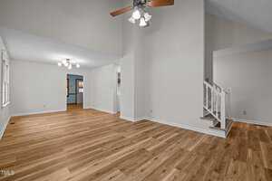 Spacious empty room with wooden flooring and light beige walls. Ceiling fans hang from the ceiling; stairs with white banisters lead to an upper level. Large windows allow natural light, and a doorway connects to another room in the background.