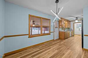 A room with light blue walls and wood flooring features a modern light fixture. There are wooden cabinets and a white door with a window. Outside the window, a red barn is partially visible. Ceiling fans are seen in both the kitchen and adjacent room.