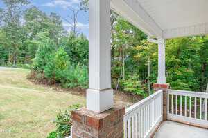A view from a porch with brick columns and white railings, overlooking a grassy yard and a wooded area. The sky is partly cloudy, and the scene is tranquil and green.