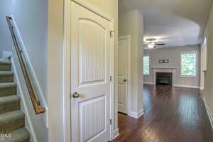 A hallway features a carpeted staircase on the left and a closed door. The hallway leads to a living room with hardwood floors, a ceiling fan, and a fireplace framed by two windows. Walls are painted a light color, and natural light fills the space.