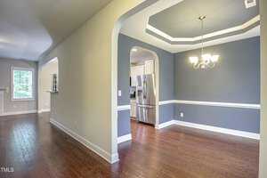 A hallway with wooden flooring leads to a room with gray walls and a white trim ceiling. A chandelier hangs above. An open doorway reveals a kitchen with white cabinets and stainless steel appliances. Natural light enters from a window.
