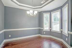 An empty room with gray walls and a dark wooden floor. A chandelier hangs from the ceiling. Wainscoting wraps around the walls. Three large windows allow natural light to enter the room.