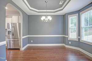 A dining room with light gray walls featuring white trim and crown molding. A silver chandelier hangs from a tray ceiling. The room has wooden floors and windows with blinds. A stainless steel refrigerator is visible through an arched doorway.