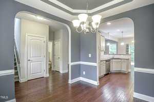A modern interior features a dining area with a chandelier and dark hardwood floors. Archways lead to the kitchen with white cabinets and stainless steel appliances. A staircase is visible on the left, and a doorway opens to another room.