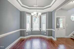 A dining room with hardwood floors, gray walls with white trim, and a chair rail. Three tall windows with blinds are on the far wall. A chandelier hangs from a tray ceiling, and a door with a curtain is to the right. Stairs are partially visible.