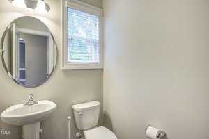A small bathroom with light-colored walls features a round mirror above a white pedestal sink. Next to the sink is a white toilet with a roll of toilet paper on a holder. A window with blinds is above the toilet, allowing natural light in.